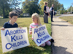 Lucy Spaetti, seated in the grass, talks with a friend while her parents Dawn and Dr. Adam Spaetti stand behind her. The family participated in the Life Chain event in front of St. Charles Borromeo Church in Bloomington on Oct. 4. (Submitted photo by Marian Leahy)